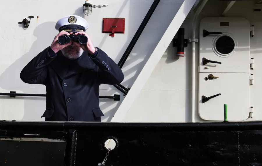 Mann in Uniform und Mütze schaut mit Fernglas von einem Schiff.