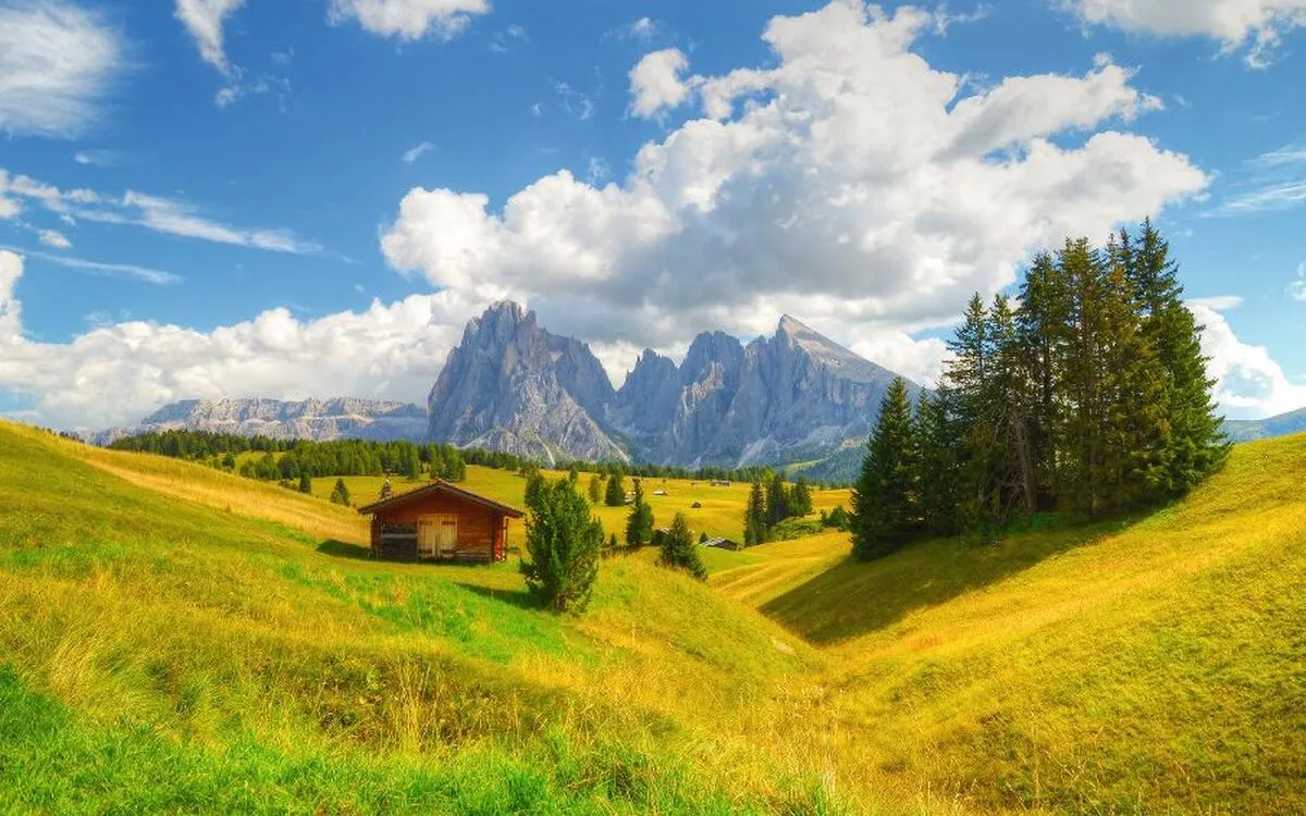 Berghütte auf einer grünen Wiese mit Gebirge im Hintergrund unter blauem Himmel.