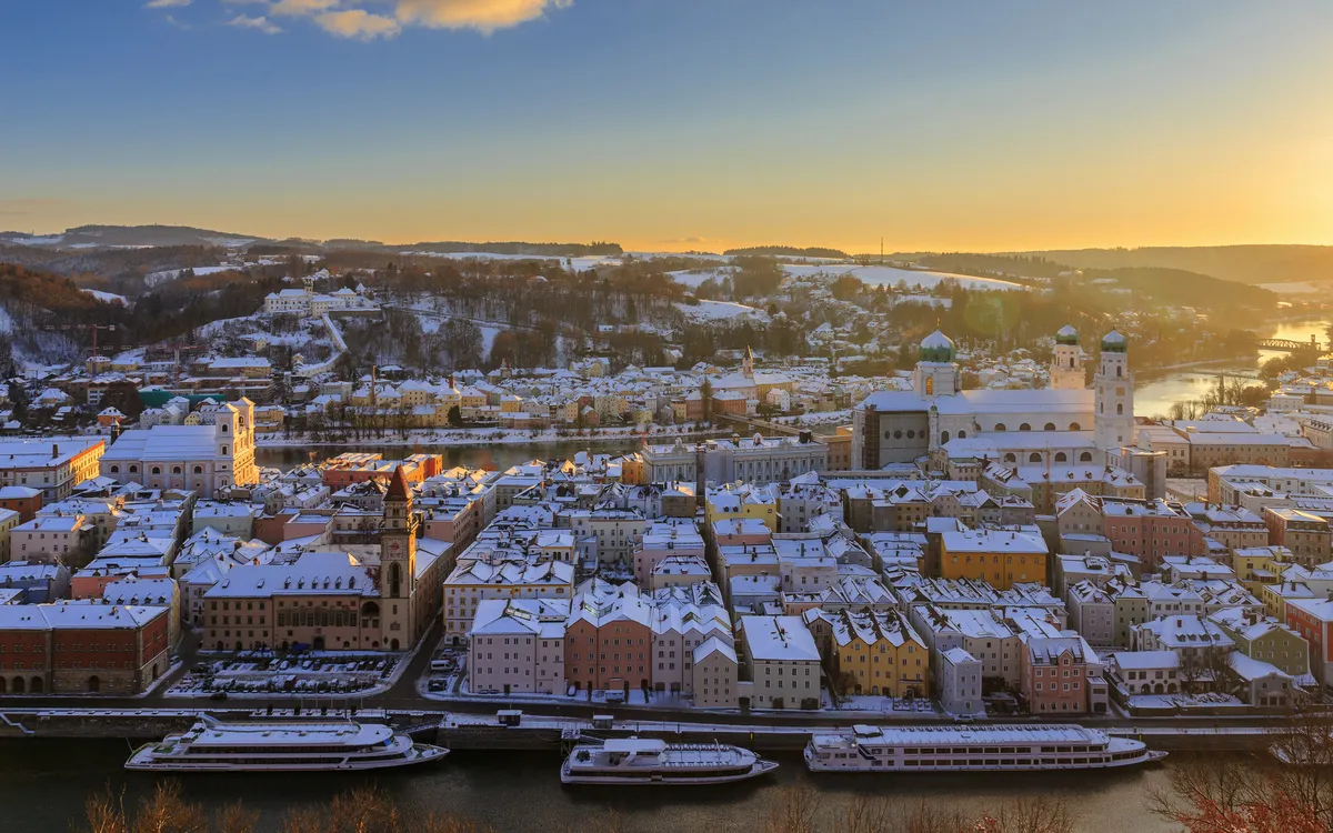 Verschneite Stadt an Flussufer bei Sonnenaufgang mit historischen Gebäuden.