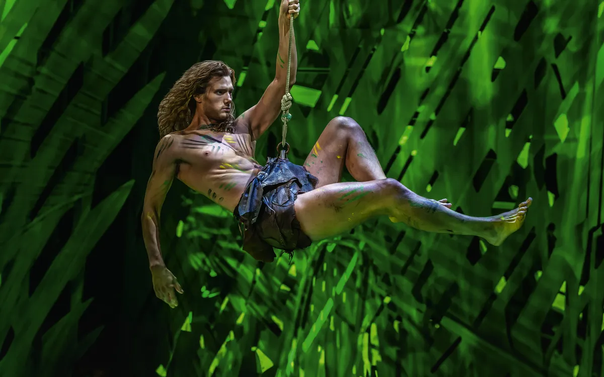 Mann in Tarzan-Kostüm schwingt an einem Seil vor grünem Hintergrund.