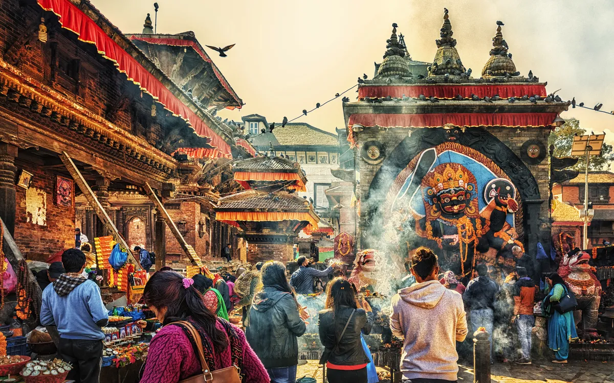 Menschen auf belebtem Markt vor traditioneller Architektur und großer Maske in Nepal.