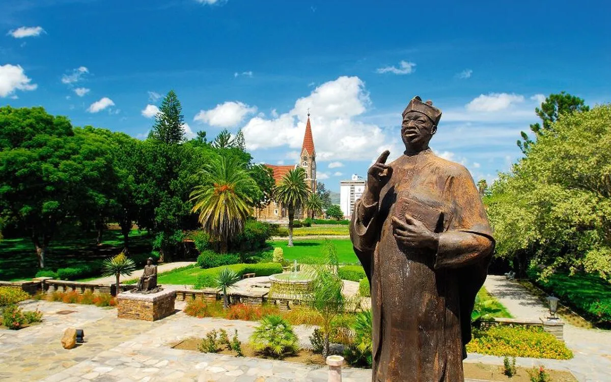 Statue im Vordergrund, Park mit Bäumen und Kirche im Hintergrund, sonniger Himmel.