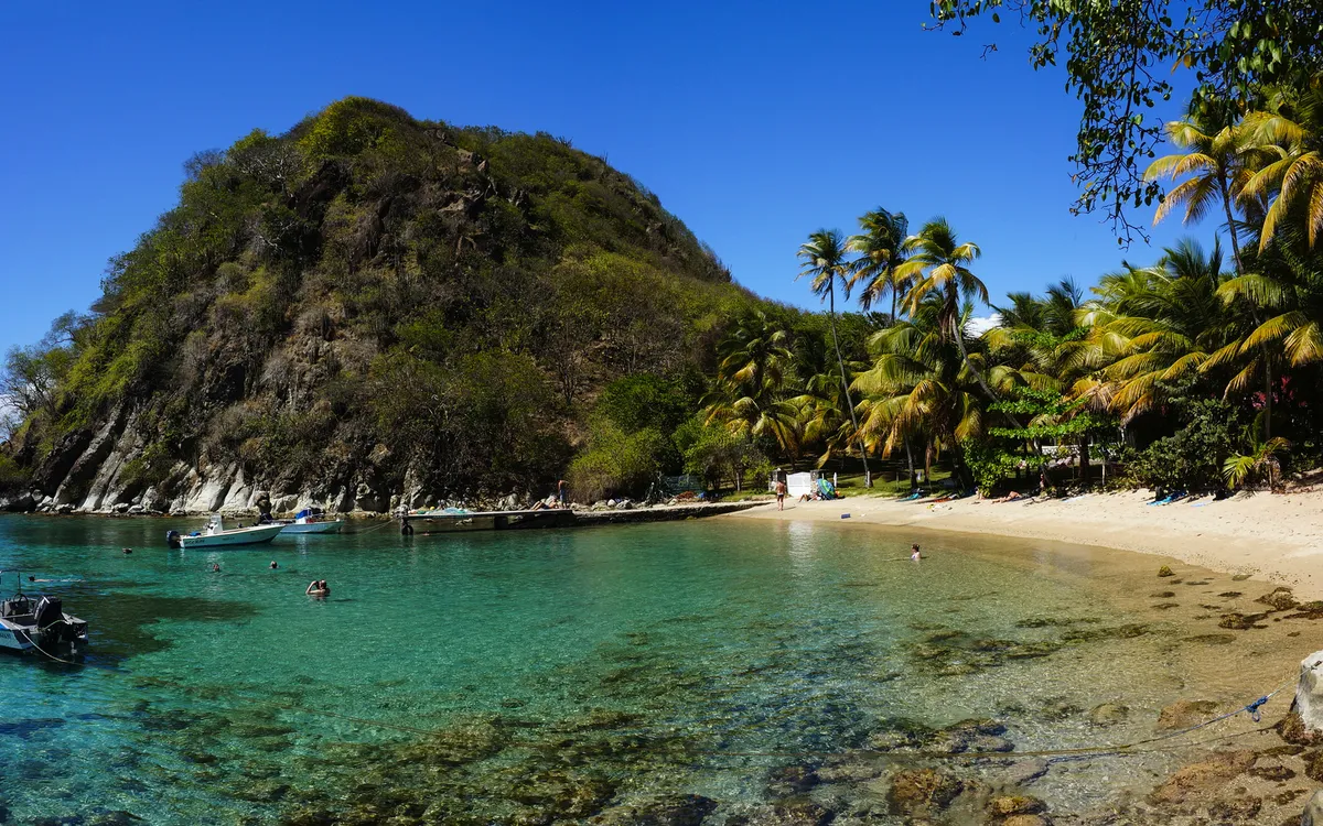 Tropischer Strand mit Palmen und klarem Wasser, Boote an der Küste, Hügel im Hintergrund.