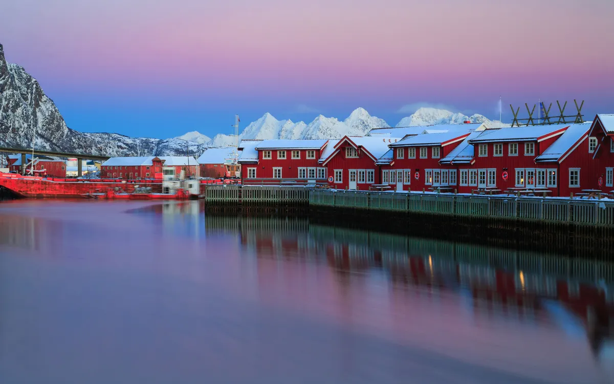 Rote Küstenhäuser vor schneebedeckten Bergen im Abendlicht.