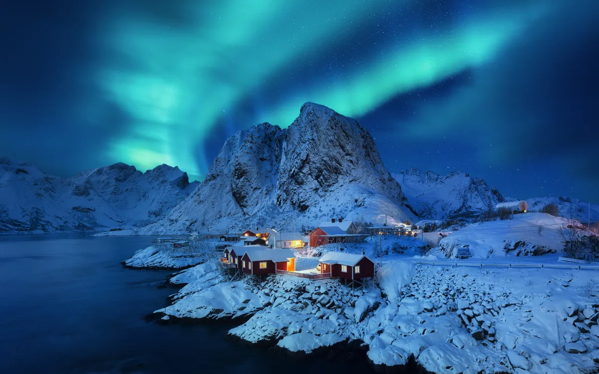 Nordlichter über einer verschneiten norwegischen Küstenlandschaft mit roten Häusern.