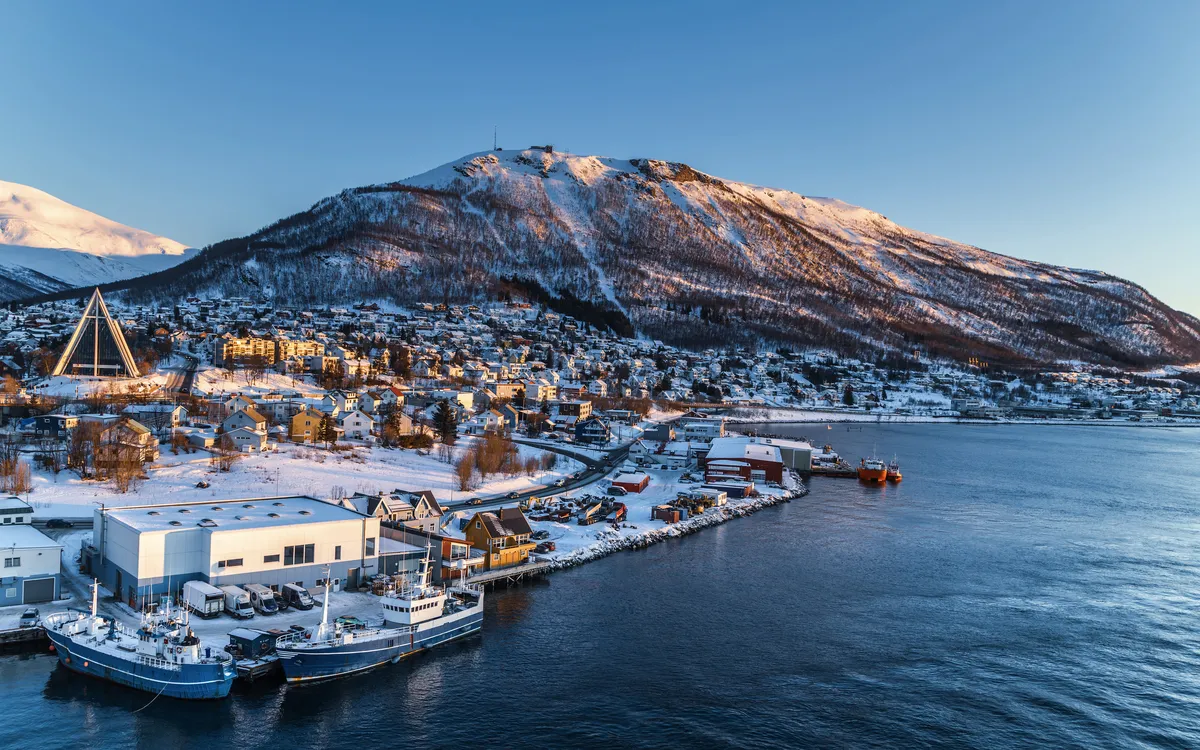 Verschneite Küstenstadt mit Hafen und Bergen im Hintergrund bei Sonnenuntergang.