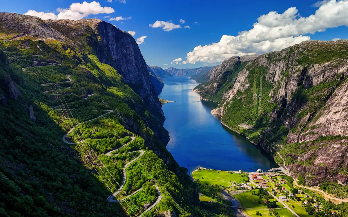 Blick auf einen Fjord mit steilen Bergen und einer kurvenreichen Straße bei sonnigem Wetter.