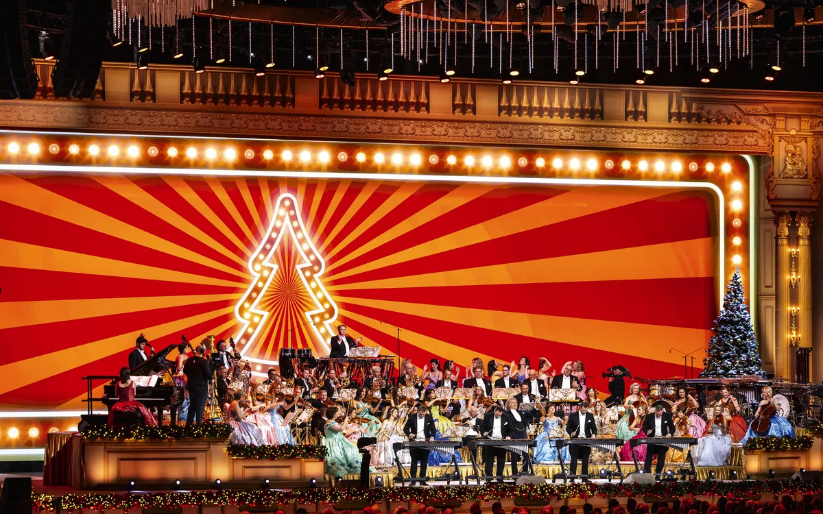 Ein Orchester spielt auf einer Bühne mit festlicher Dekoration und leuchtendem Hintergrund.
