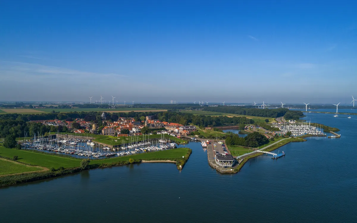 Luftaufnahme einer Küstenstadt mit Yachthafen und Windrädern im Hintergrund.