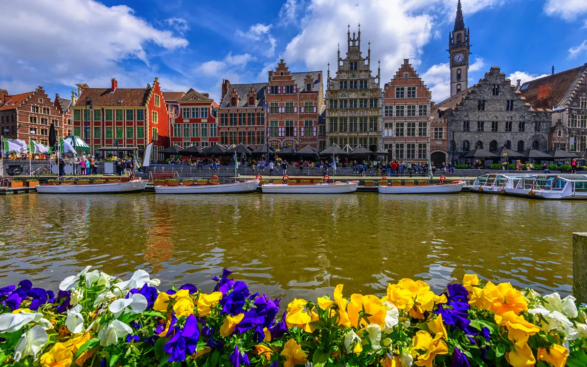 Blumenbeet vor bunten Häusern und Kanal in einer historischen Stadt bei klarem Himmel.