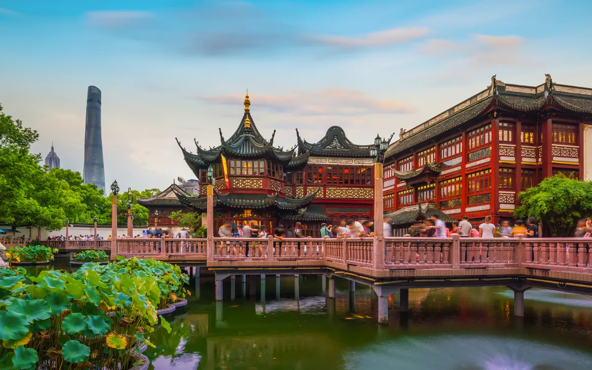 Traditionelle chinesische Architektur mit Brücke über Teich und modernem Turm im Hintergrund.
