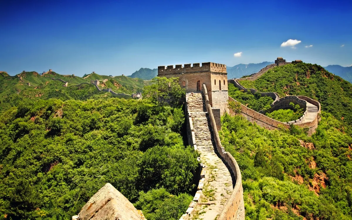 Chinesische Mauer schlängelt sich durch grüne hügelige Landschaft unter blauem Himmel.