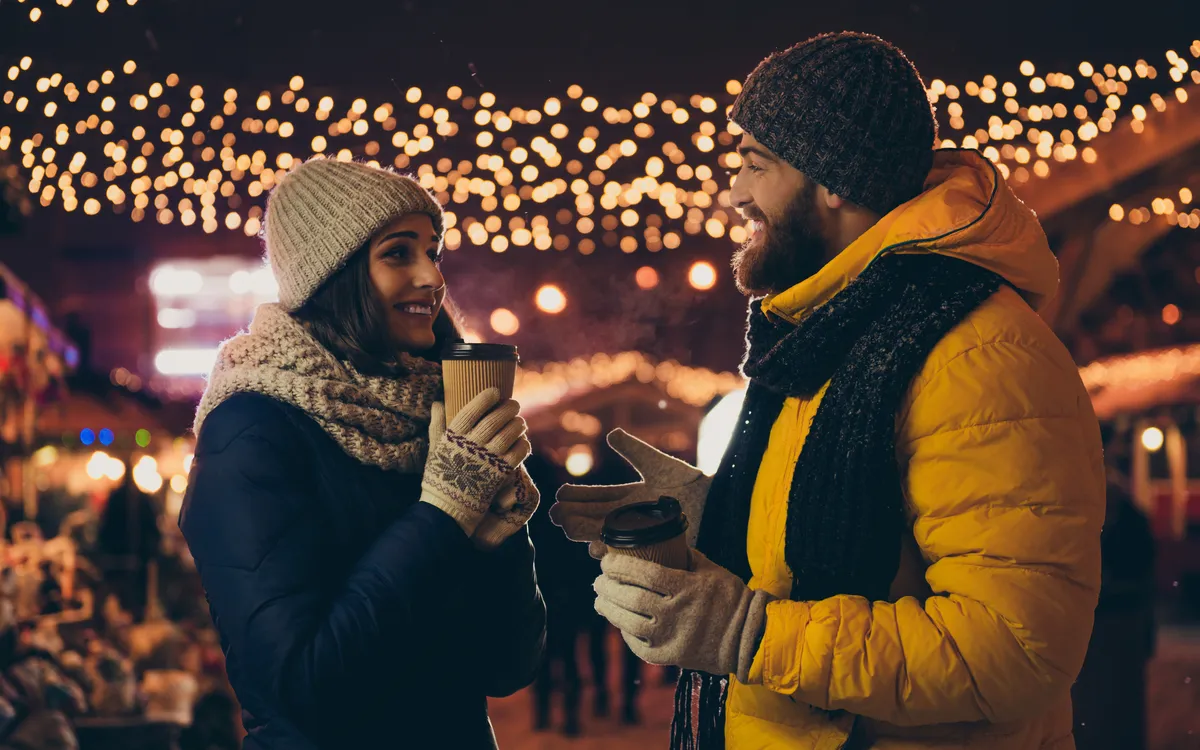 Paar mit warmen Getränken auf einem festlichen Weihnachtsmarkt bei Nacht.