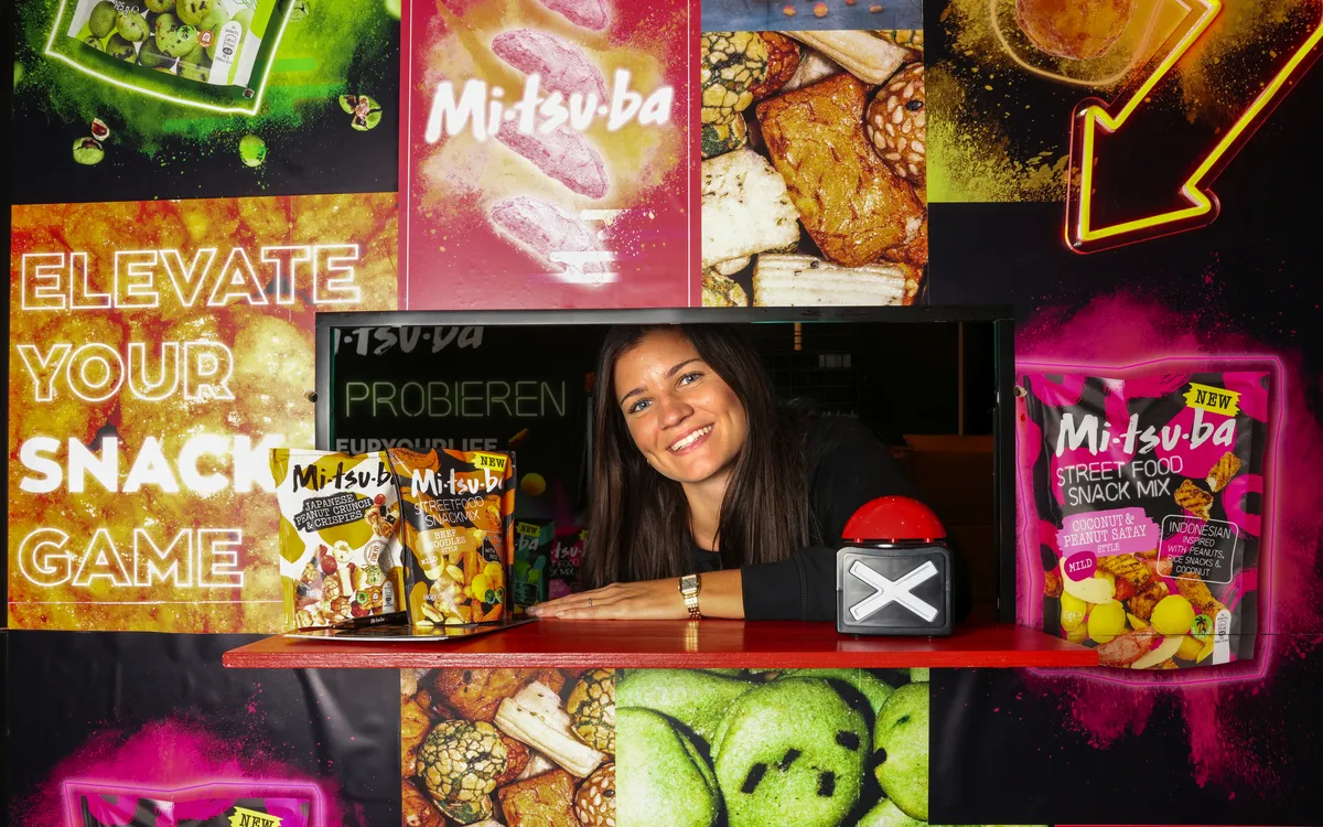 Frau präsentiert Snacks vor farbenfrohem Werbeaufsteller.