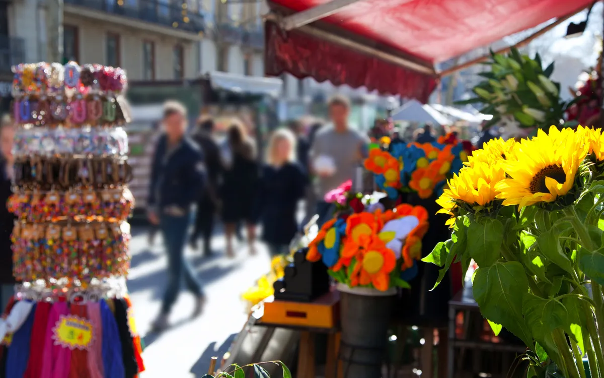 Marktstand mit Blumen und Schmuck, unscharfe Passanten im Hintergrund.