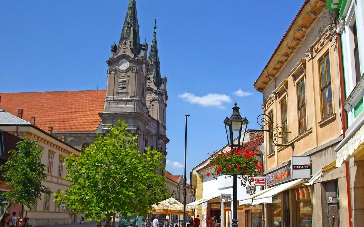 Straßenszene mit historischer Kirche und Geschäften bei sonnigem Wetter.