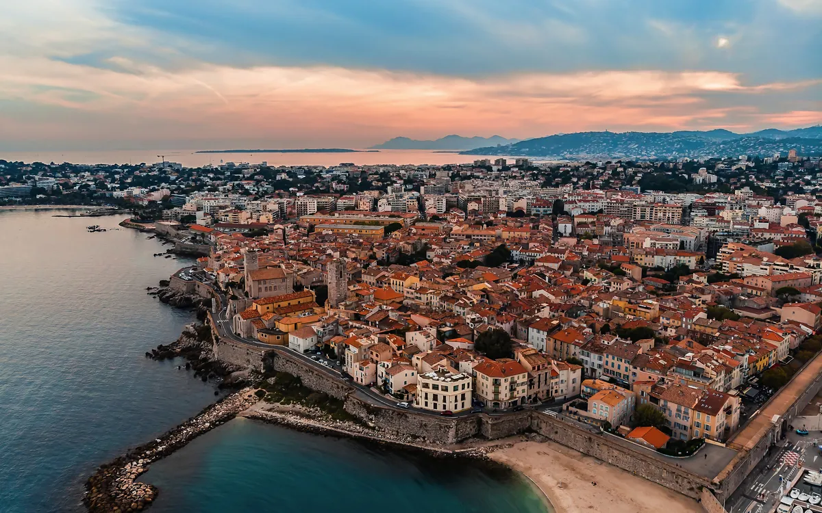 Luftaufnahme einer Küstenstadt mit historischen Gebäuden und Sandstrand bei Sonnenuntergang.
