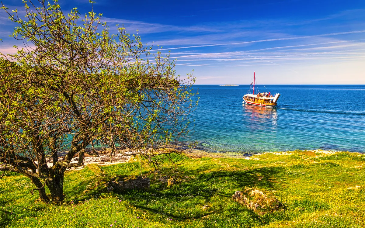 Segelboot auf türkisfarbenem Meer, grüner Wiese im Vordergrund, blauem Himmel im Hintergrund.