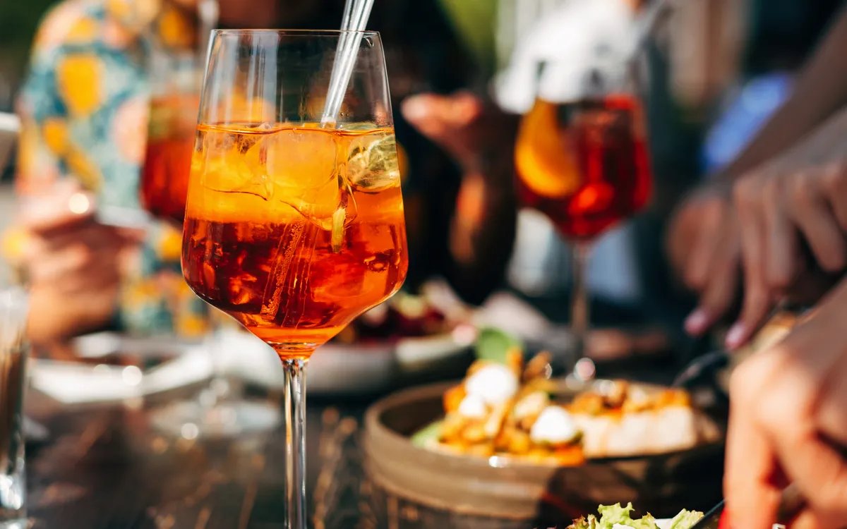 Sommer-Cocktail Aperol Spritz auf einem Esstisch, umgeben von Menschen, die Speisen genießen.