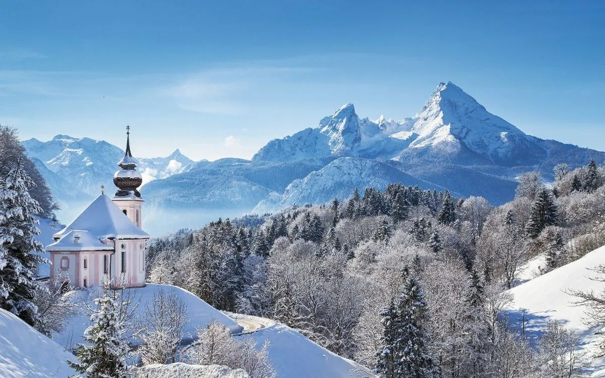 Winterlandschaft mit einer kleinen Kirche im Vordergrund und schneebedeckten Bergen im Hintergrund.