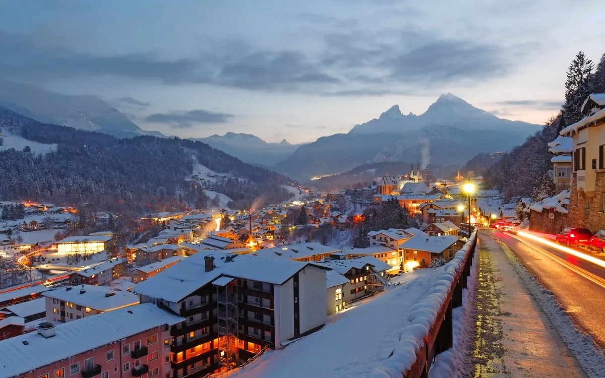 Verschneite Stadtlandschaft am Abend mit leuchtenden Häusern und Bergen im Hintergrund.