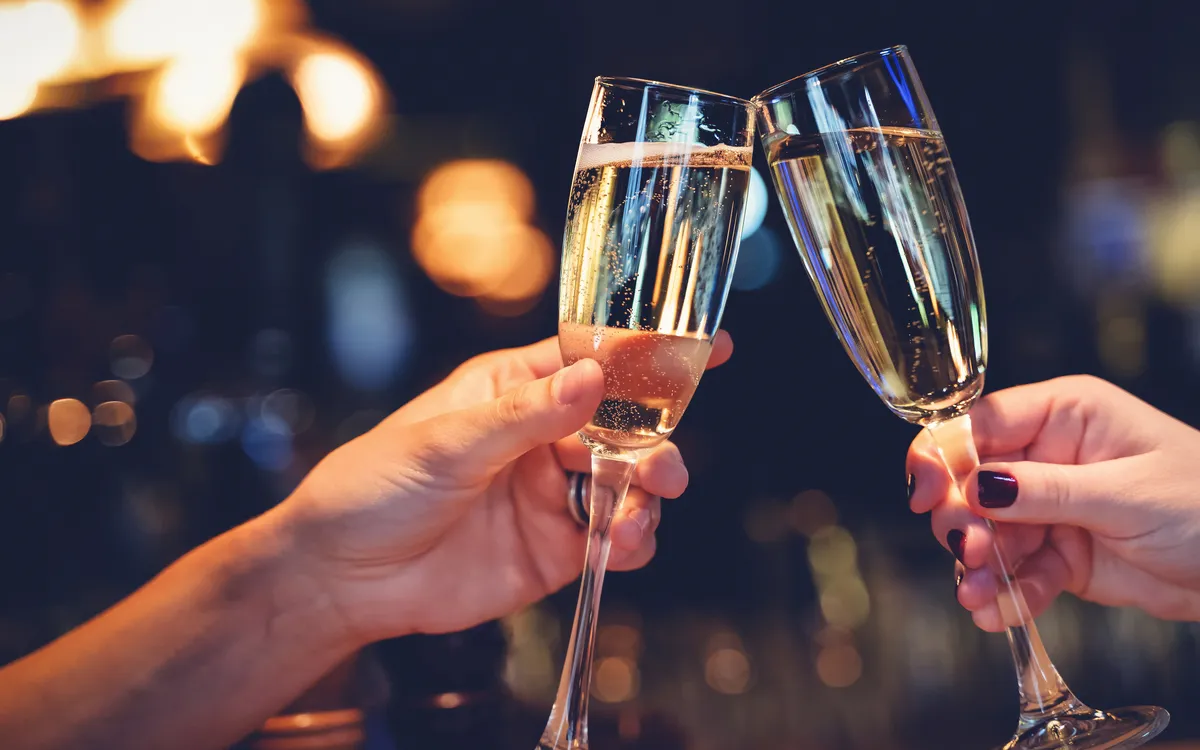 Zwei Personen stoßen mit Champagnergläsern an, unscharfer Hintergrund mit warmer Beleuchtung.