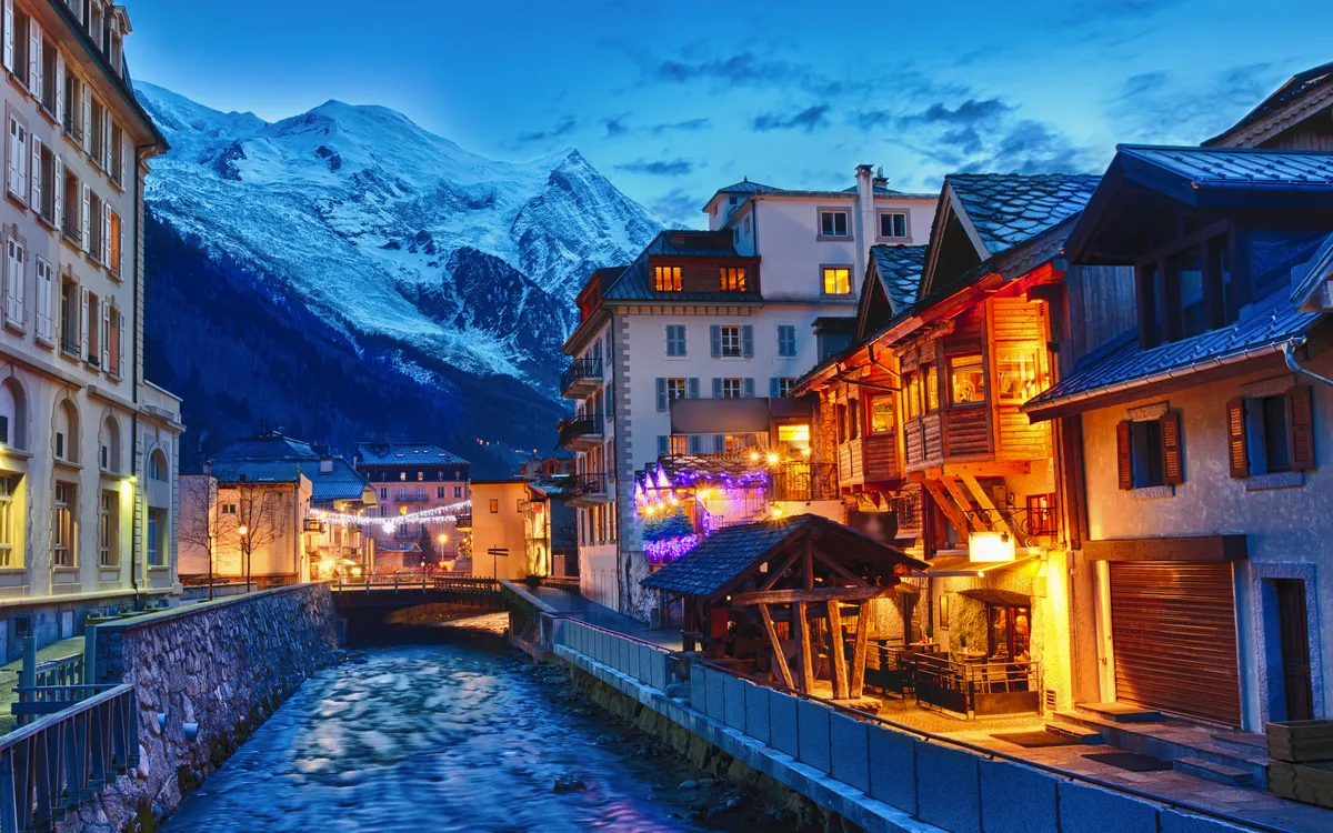 Bergdorf mit Fluss, beleuchteten Häusern und schneebedeckten Bergen im Hintergrund.