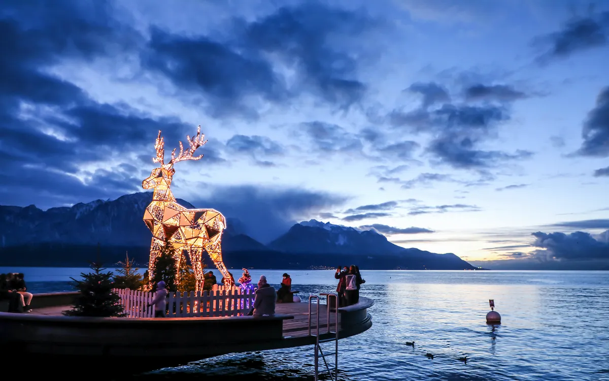 Beleuchtete Hirschstatue am Seeufer bei Sonnenuntergang mit Bergen im Hintergrund.