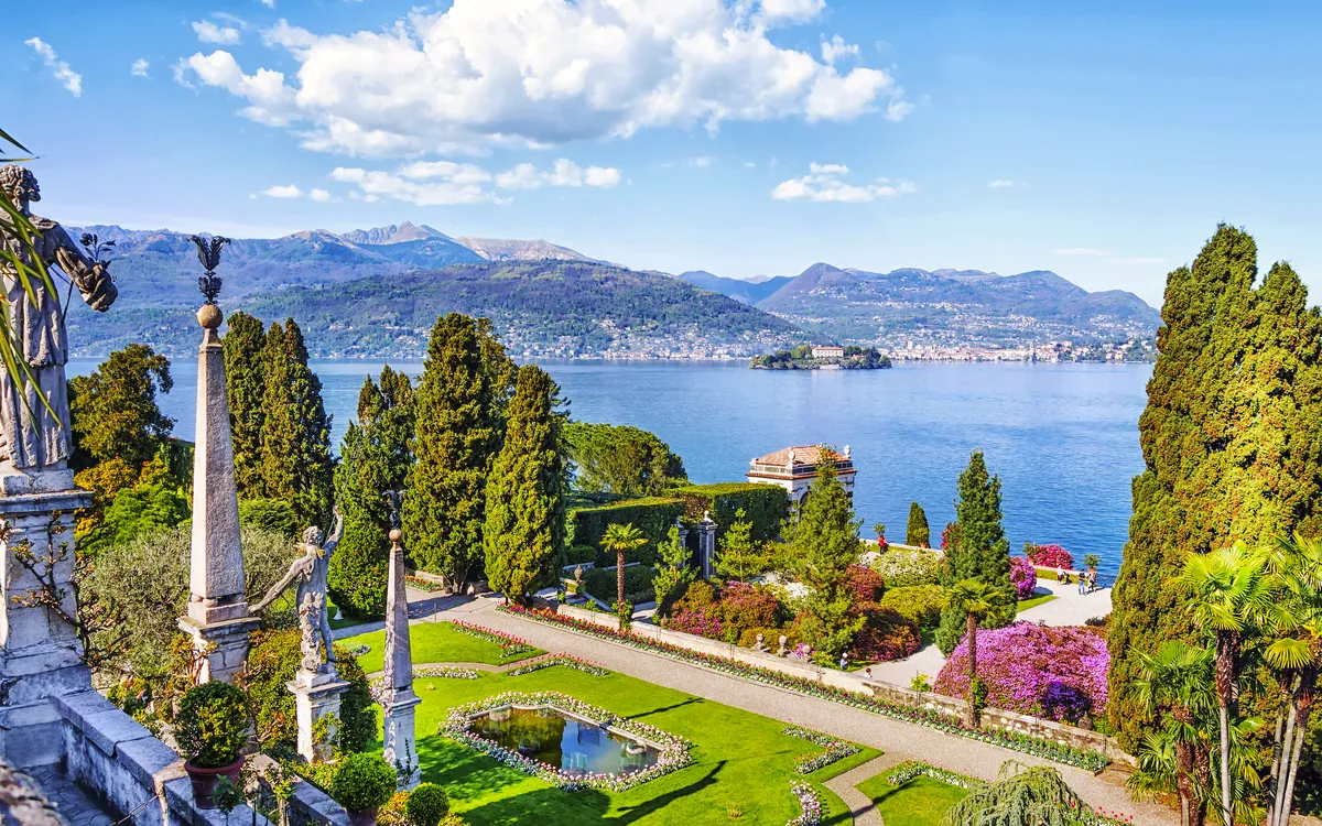 Garten mit Springbrunnen und Bäumen am Seeufer mit Bergen im Hintergrund.