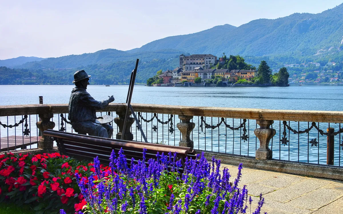 Statue eines Malers am Seeufer mit Blick auf eine Insel und Berge im Hintergrund.
