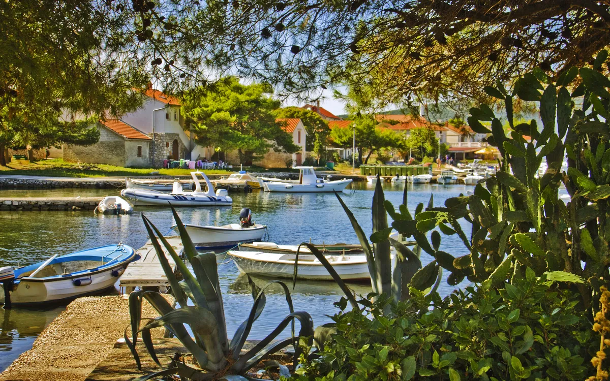 Boote in einem kleinen Hafen, umgeben von mediterranen Pflanzen und Häusern mit roten Dächern.