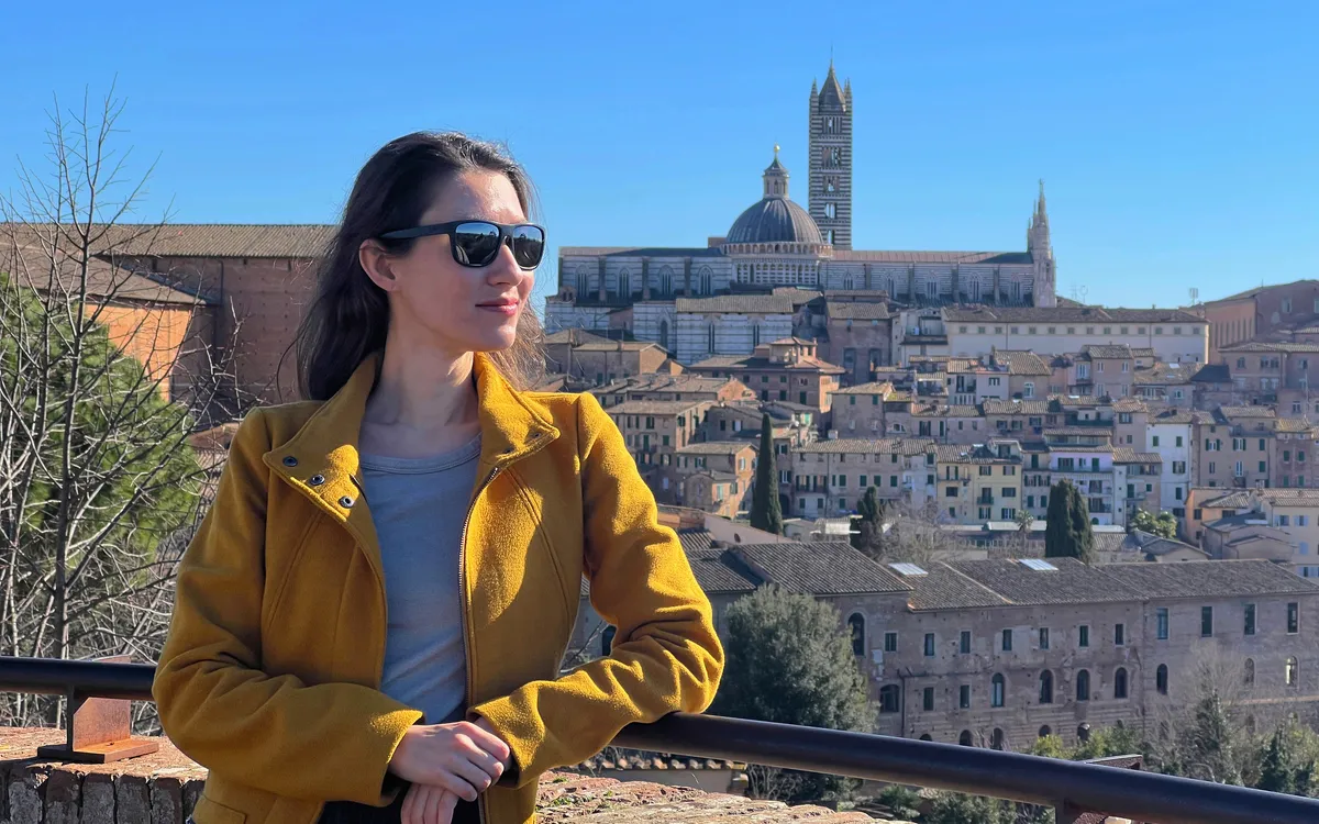 Eine Frau in gelber Jacke mit Sonnenbrille vor Stadtansicht von Siena.