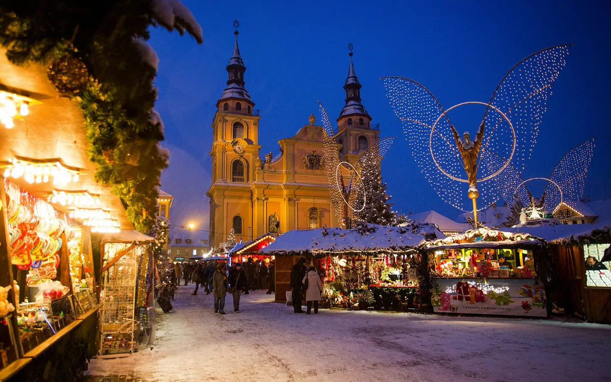 Weihnachtsmarkt mit Ständen, Kirche und großer Engel-Lichtinstallation bei Schnee.