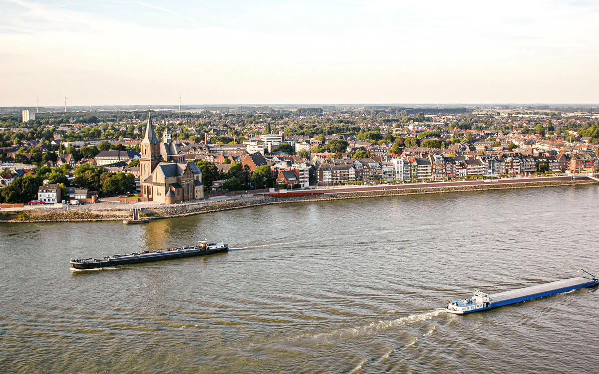 Zwei Frachtschiffe fahren auf einem Fluss vor einer Stadt mit Kirche im Hintergrund.