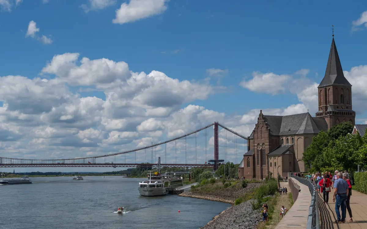 Kirche am Fluss mit Menschen und Brücke im Hintergrund bei blauem Himmel