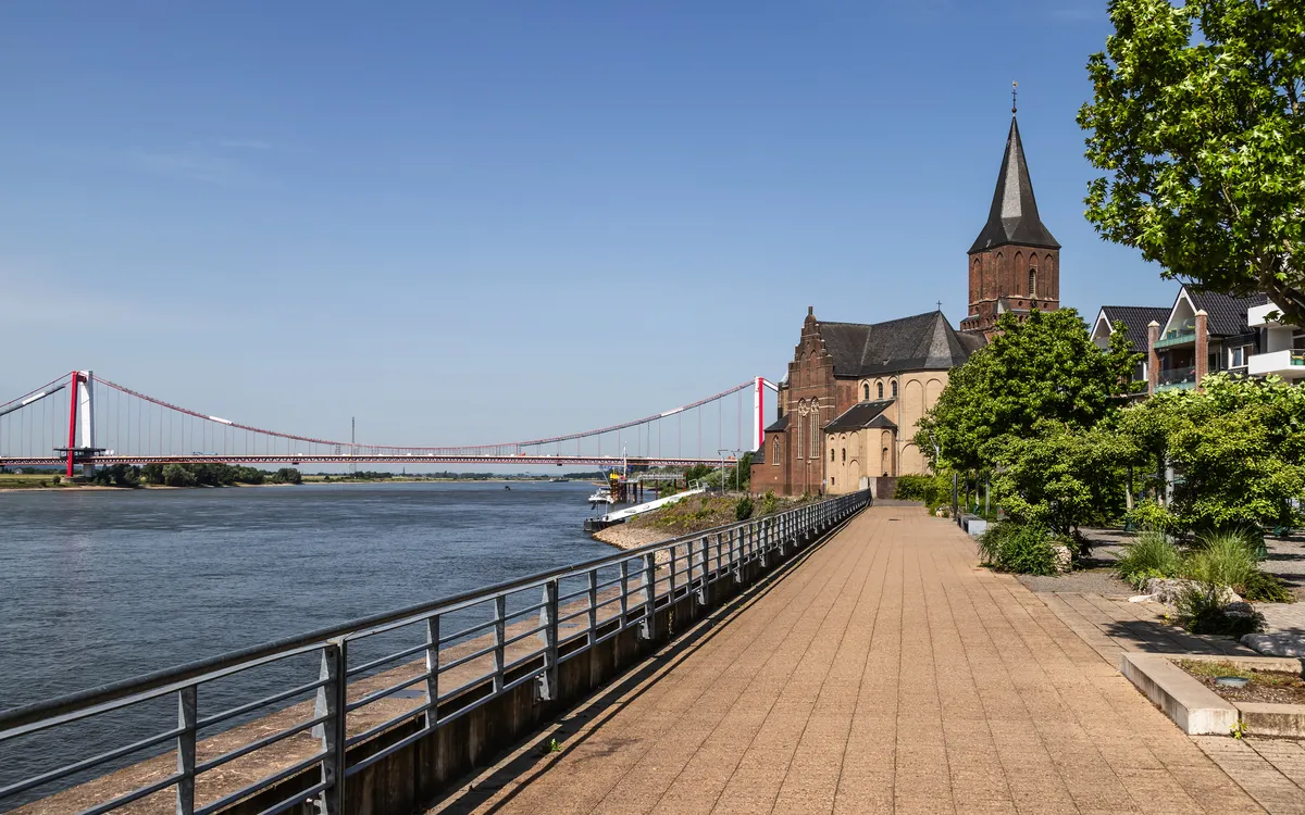 Rheinpromenade mit Kirche und Brücke in der Ferne bei klarem Himmel.