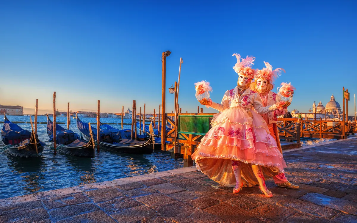 Zwei Personen in venezianischen Karnevalskostümen posieren am Wasser bei Sonnenuntergang.