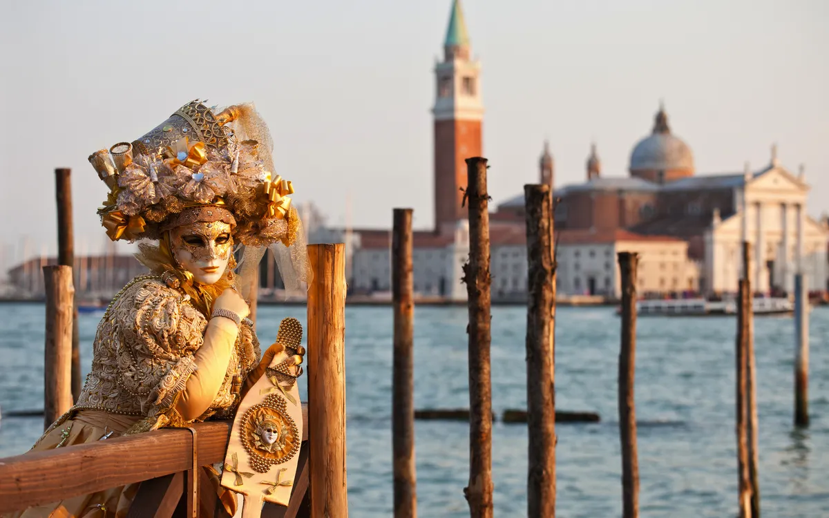 Person in venezianischem Karnevalskostüm vor Wasser und Kirche in Venedig.