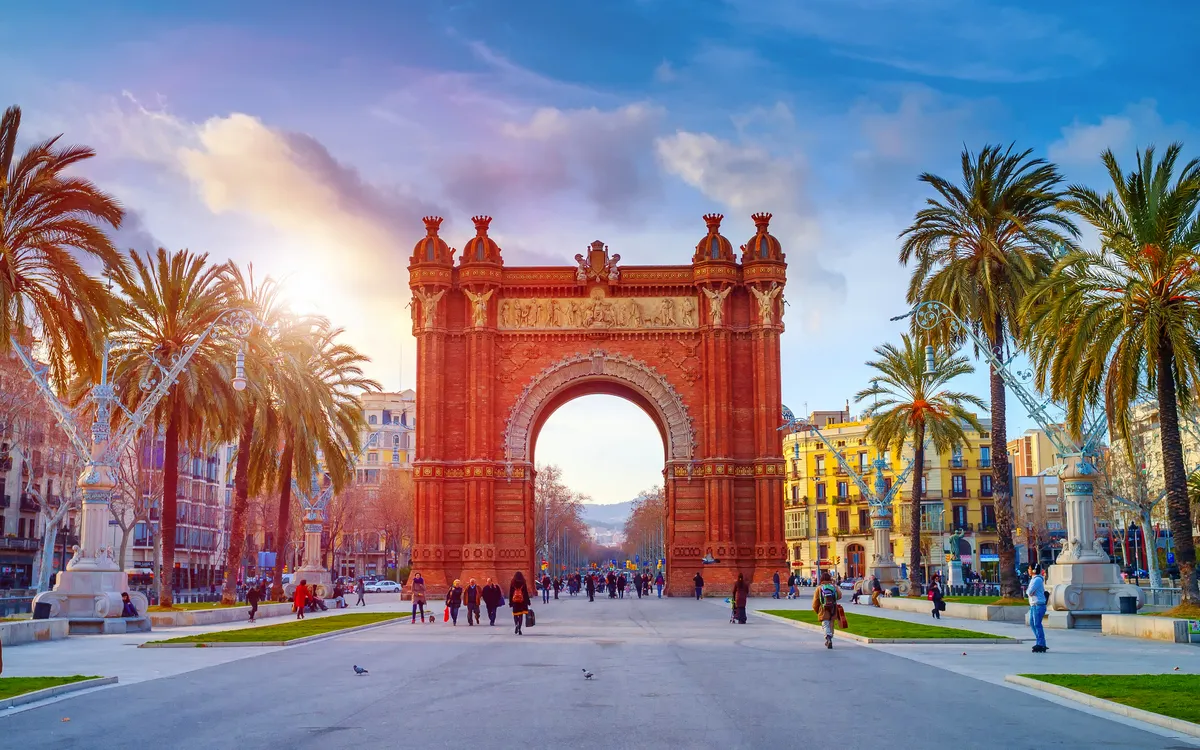 Triumphbogen in Barcelona bei Sonnenschein mit Menschen und Palmen umgeben.