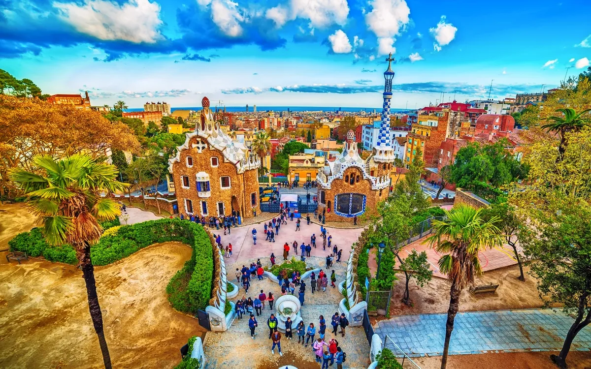 Menschenmenge im Park Güell in Barcelona mit Blick auf bunte Gebäude und die Stadt im Hintergrund.