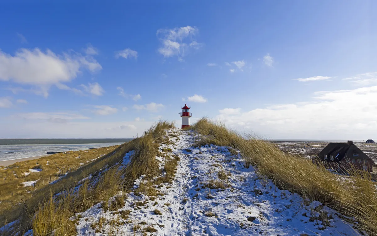 Leuchtturm auf einer verschneiten Dünenlandschaft am Meer bei blauem Himmel.