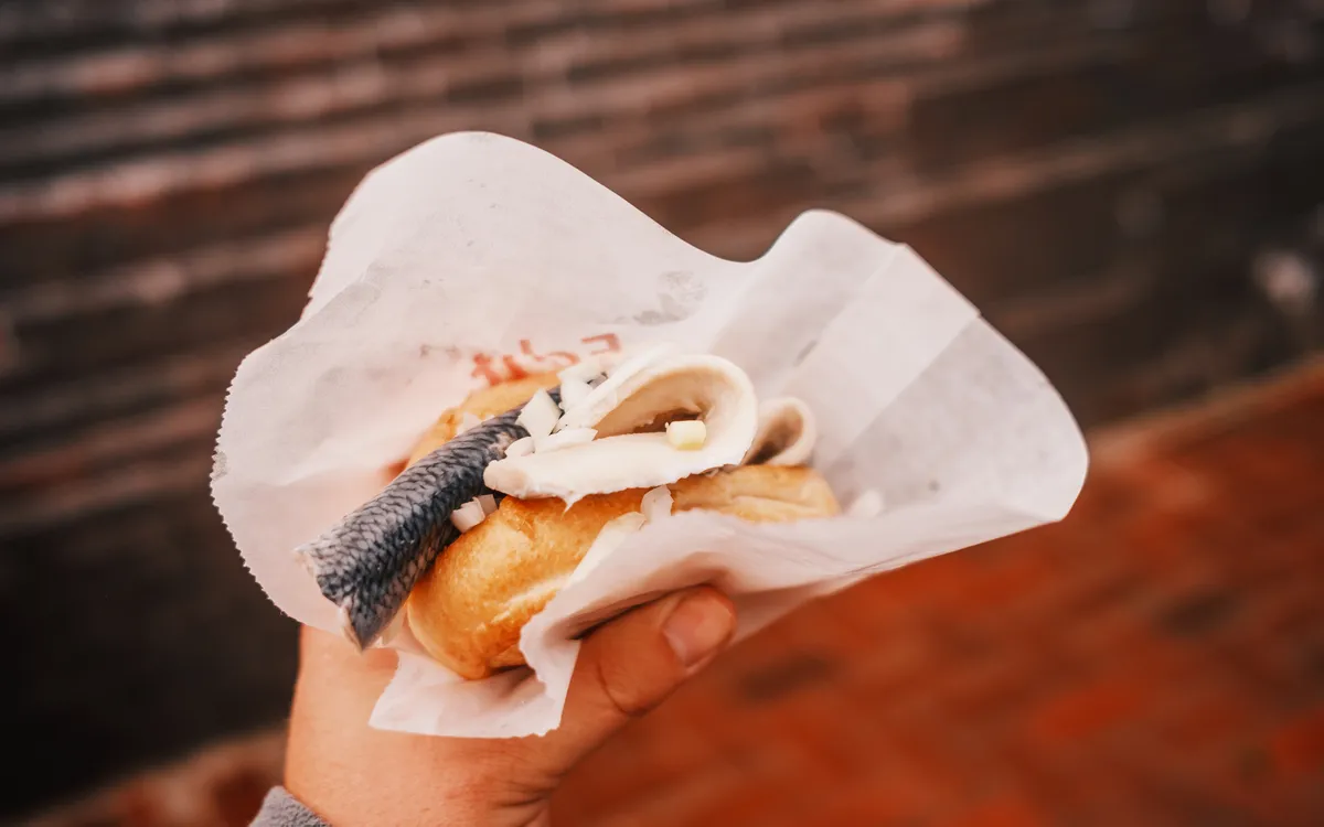 Hand hält Fischbrötchen in Papierverpackung vor einer Ziegelmauer.
