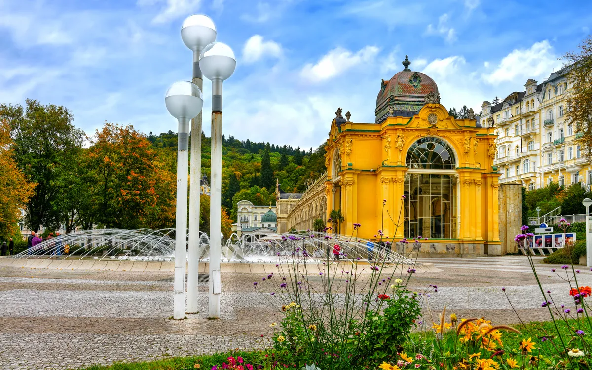 Gelbes historisches Gebäude mit Springbrunnen im Vordergrund und blühende Pflanzen.