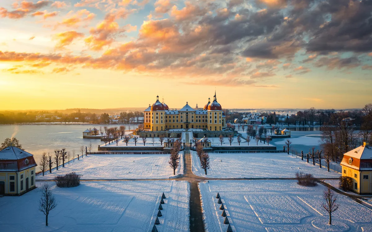 Schloss im Winter bei Sonnenuntergang, umgeben von verschneiten Gärten und einem See.