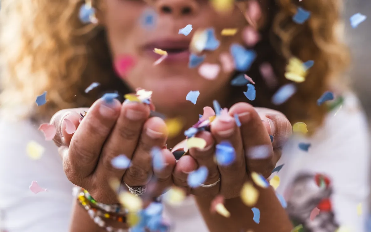 Frau in ihren 40ern bläst buntes Konfetti aus den Händen, Party-Atmosphäre.