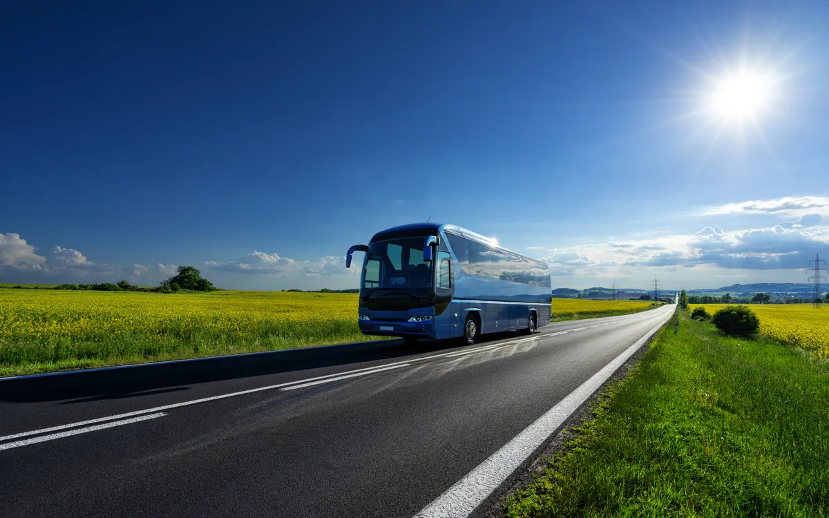Reisebus fährt bei Sonnenschein auf einer Asphaltstraße durch Rapsfelder.