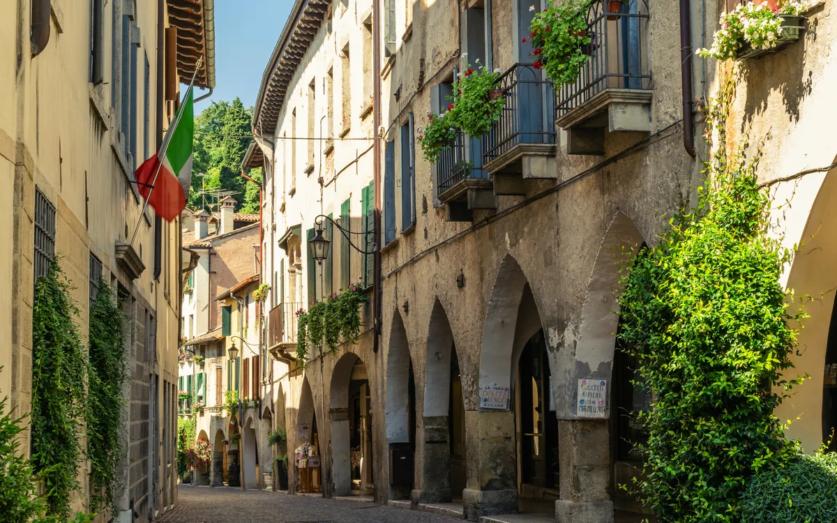 Schmale, gepflasterte Straße mit alten Gebäuden und italienischer Flagge.