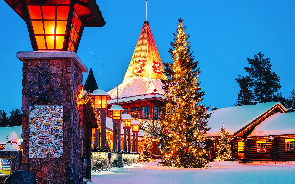 Verschneiter Weihnachtsdorfplatz mit beleuchteten Gebäuden und großem Tannenbaum.
