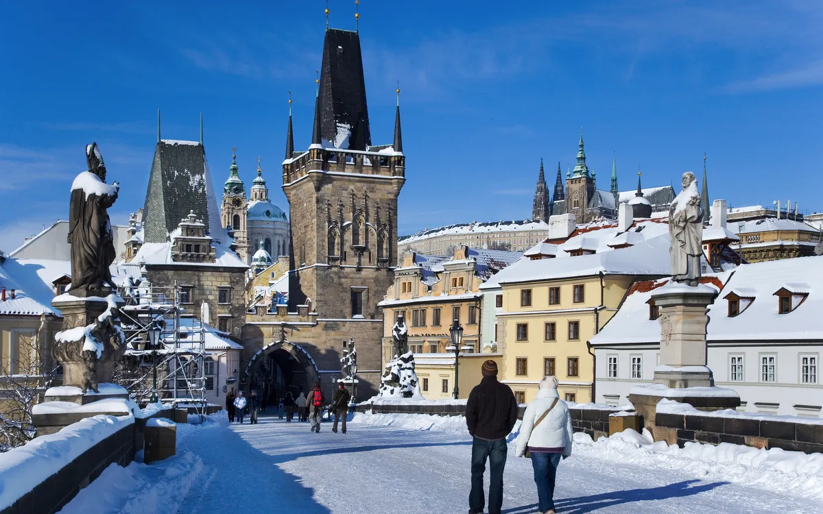 Menschen gehen im Winter über die Karlsbrücke in Prag, im Hintergrund historische Gebäude.