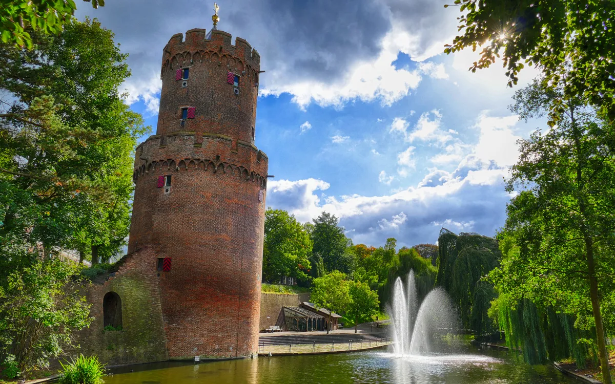 Runder Backsteinturm am See mit Springbrunnen und Bäumen im Hintergrund.